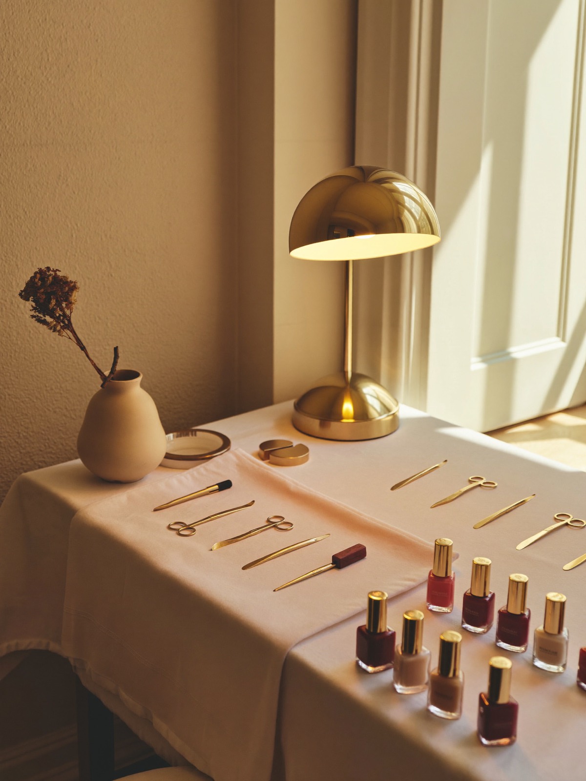Sfeerbeeld van het atelier: manicuretafel met gouden lamp, tools en nagellak in warme tinten